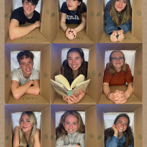 Nine students on their elbows in a cardboard box structure, smiling at the camera
